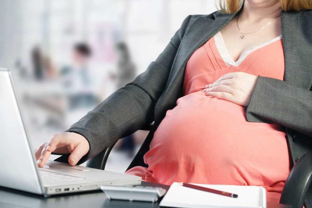Zmiany w urlopach macierzyńskich i rodzicielskich - kobieta w ciąży siedzi przy biurku i pisze na klawiaturze laptopa jedną dłonią, drugą trzyma na brzuchu. 