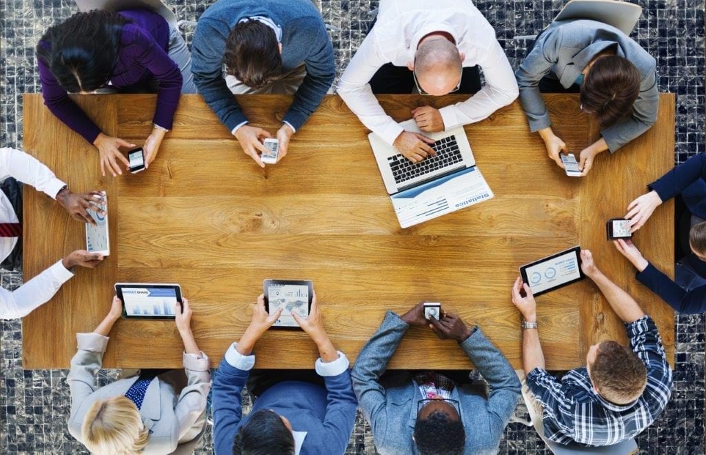 Transformacja cyfrowa w firmach przyspiesza z powodu oczekiwań klientów - ludzie siedzą przy stole i trzymają w rekach urządzenia mobilne. 