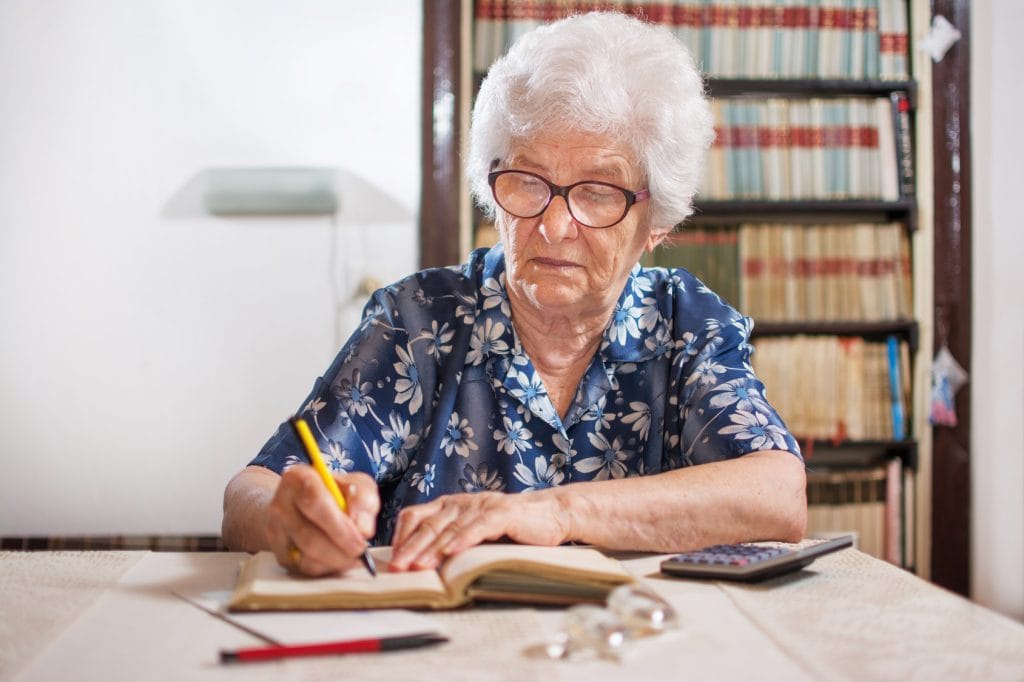 Biedny jak senior - starsza kobieta siedzi przy stole i notuje w zeszycie, obok leży kalkulator. 