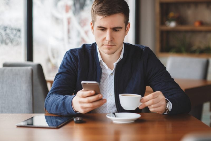Wakacje i praca w jednym – utopia czy nowa rzeczywistość? - mężczyzna pije kawę w kawiarni i trzyma w ręce telefon. 