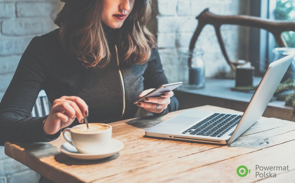 Praca zdalna 2023 - zmiany w Kodeksie pracy - kobieta siedzi przy stole z telefonem w ręku, na którym stoi laptop i kawa.