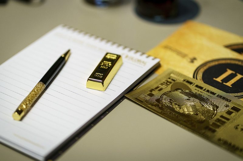 Rekordowy popyt na złoto - Polacy w 2022 kupili ponad 18 ton kruszcu - sztabka złota lezy na notesie obok banknotów i długopisu. 