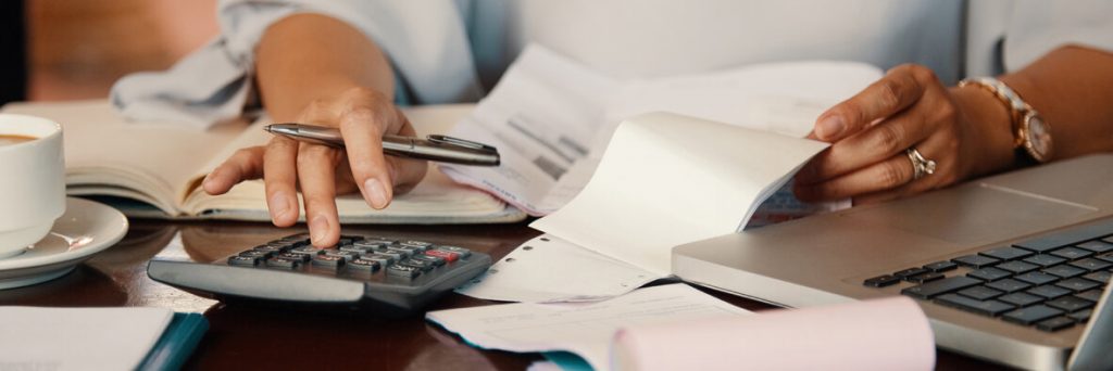 Jak niespłacone długi utrudniają codzienne życie?  - biurko z dokumentami i dłonie kobiece, które liczą na kalkulatorze.
