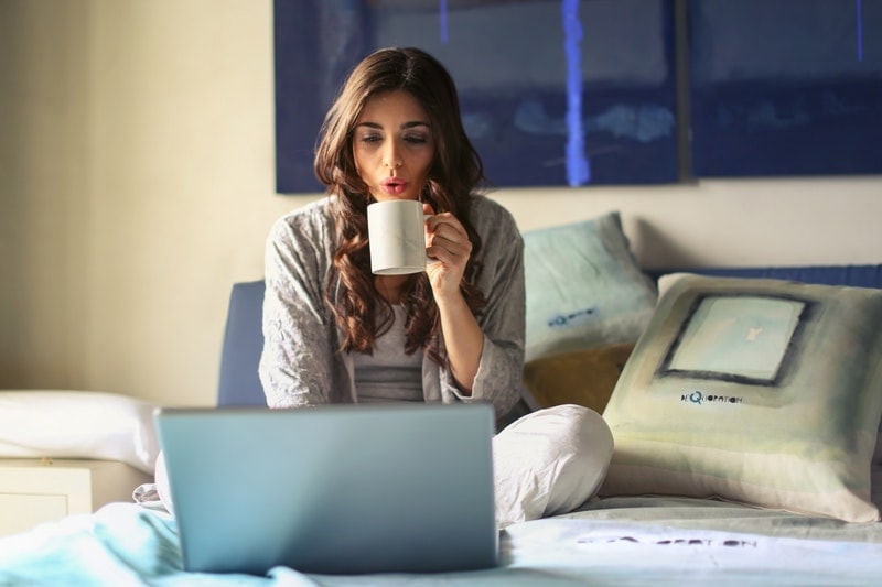 Marketing sieciowy – sposób na biznes czy zajęcie dodatkowe? - kobieta z kubkiem w reku siedzi na łóżku przed otwartym komputerem.