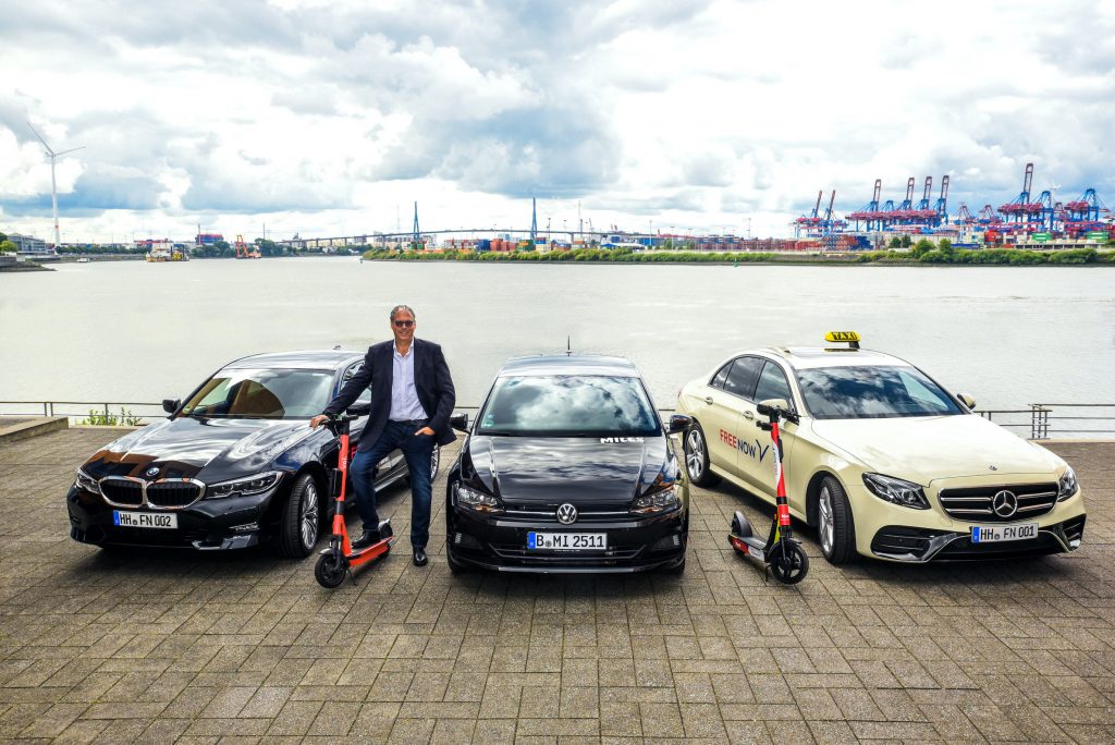 FREE NOW w Niemczech integruje carsharing - męzczyzna stoi na tle 3 aut wraz z hulajnogą