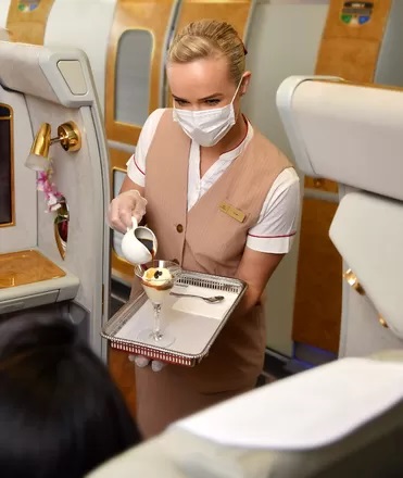Linie Emirates rozlewają 28 milionów filiżanek kawy rocznie - kobieta w maseczce na twarze rozlewa kawę na pokładzie samolotu. 