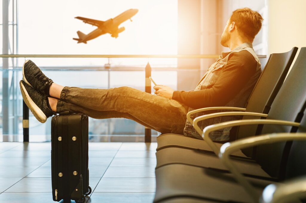 Czy bilety lotnicze będą tańsze w tym roku? - mężczyzna siedzi na lotnisku z założonymi nogami na walizce i patrzy na startujący samolot. 