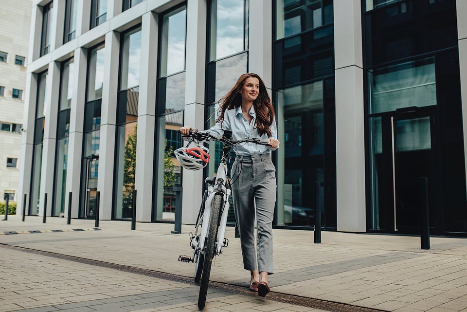 Polacy chcą rowerów elektrycznych z dofinansowaniem  - kobieta idzie obok roweru, na kierownicy zawieszony ma kask, w tle szklany budynek.