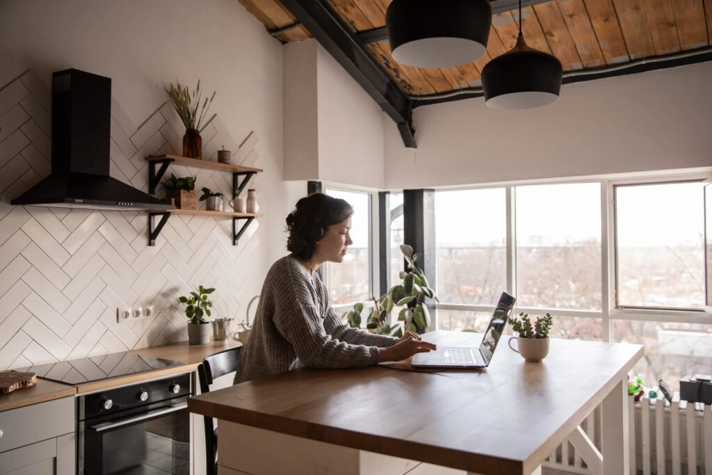 Większe mieszkanie nie oznacza większego szczęścia - kobieta w kuchni stoi przez wyspą, na której stoi laptop.