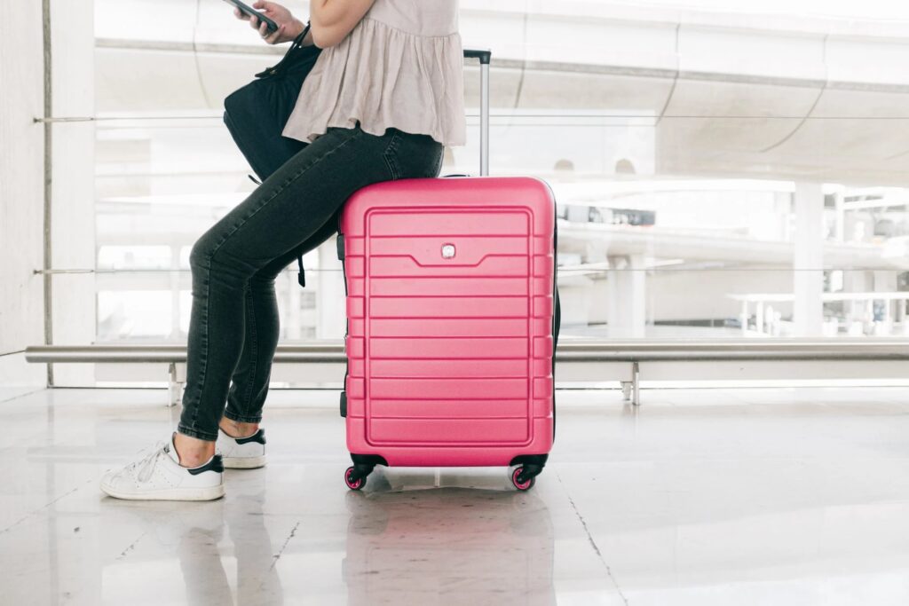 Wakacje last minute - sprawdź, na co uważać, by nie stracić pieniędzy! - kobieta siedzi na różowej walizce na lotnisku.
