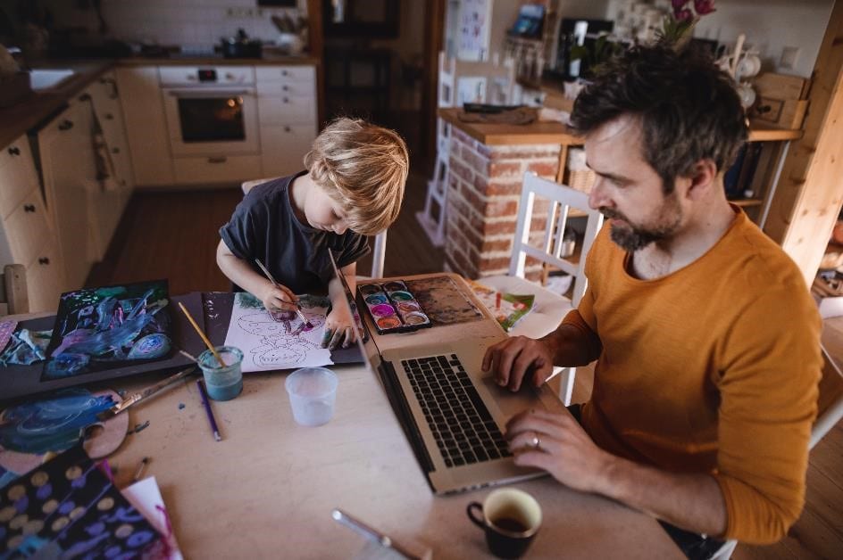 Ważne zmiany w rozliczeniach kosztów mieszkania w działalności gospodarczej - mężczyzna siedzi przy stole i pisze na klawiaturze laptopa, obok rysuje dziecko.