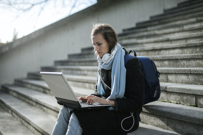 młoda kobieta w płaszczu i z plecakiem korzystająca z komputera na schodach